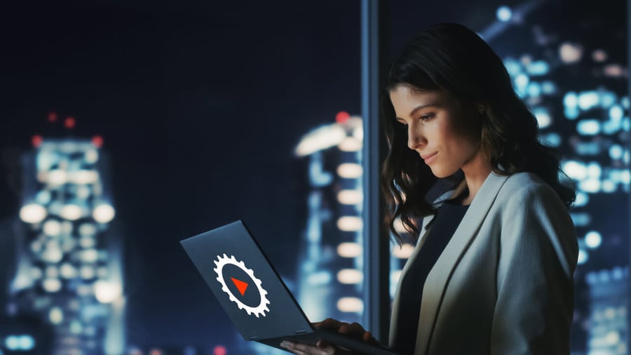 A female executive, working with their laptop in the top floor of a high rise and a nighttime city skyline is in the background