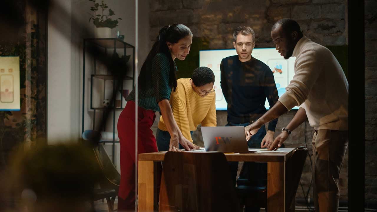 A group of diverse, professional peers in a modern office setting contemplating the companies strategy and in the foreground there's a laptop with the ReadyWorks lettermark logo